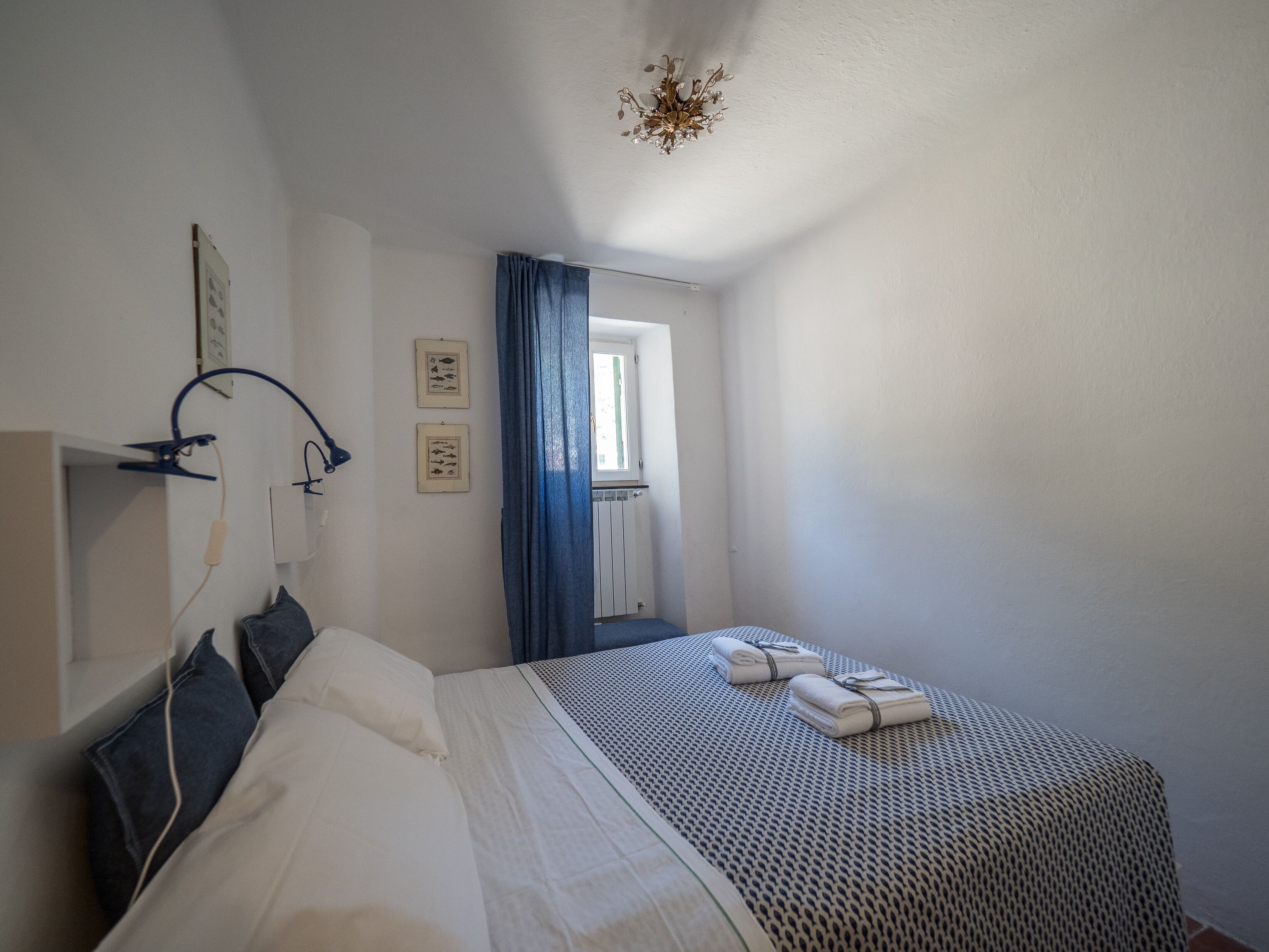 Une chambre à coucher avec un lit, une fenêtre avec des rideaux bleus, une lampe fixée au mur et un luminaire au plafond.