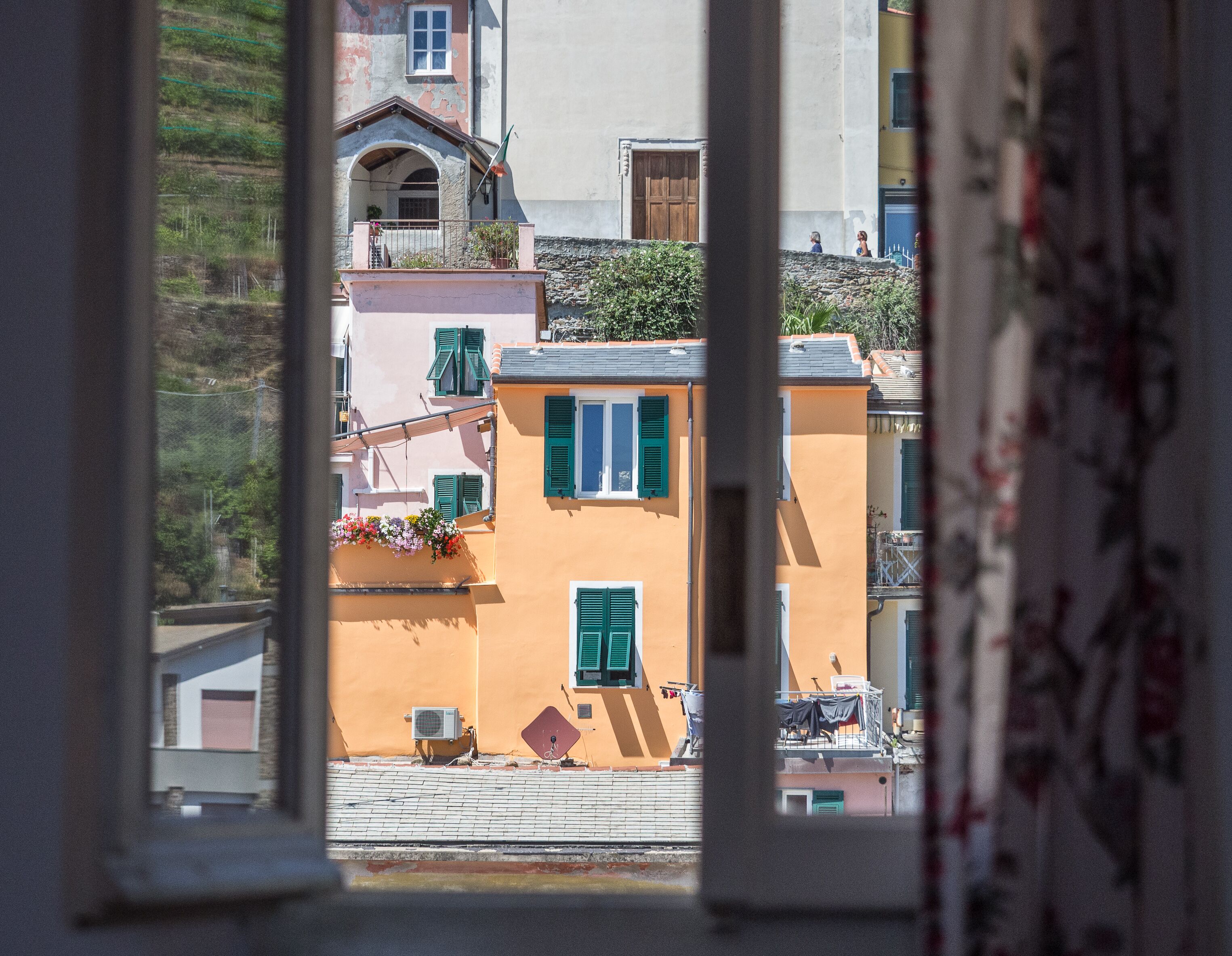 A view through a window of a building with a balcony, a small garden, and a street scene.