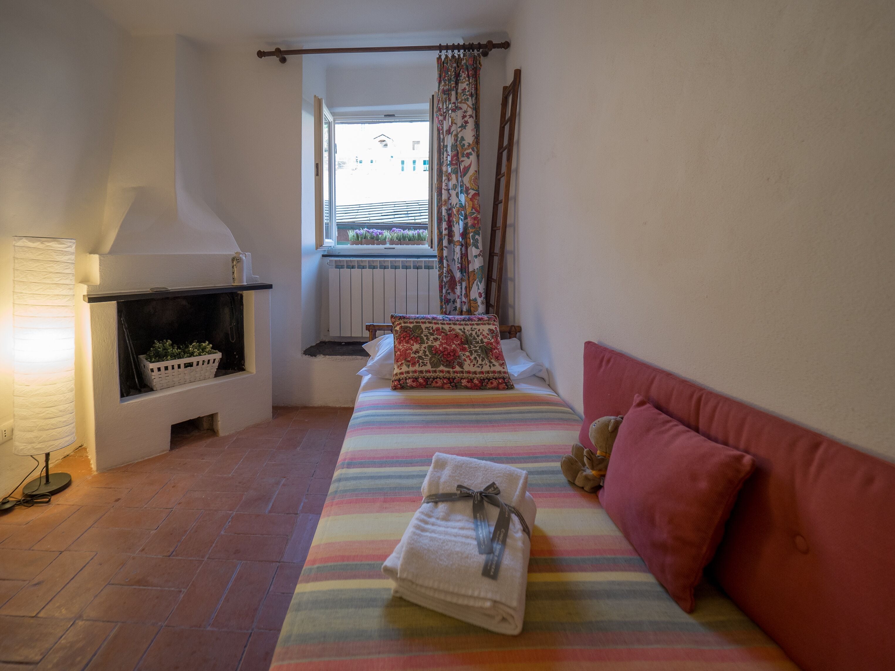 Een gezellige kamer met een open haard, een bed met een gestreept plaid en een raam met bloemen gordijnen.