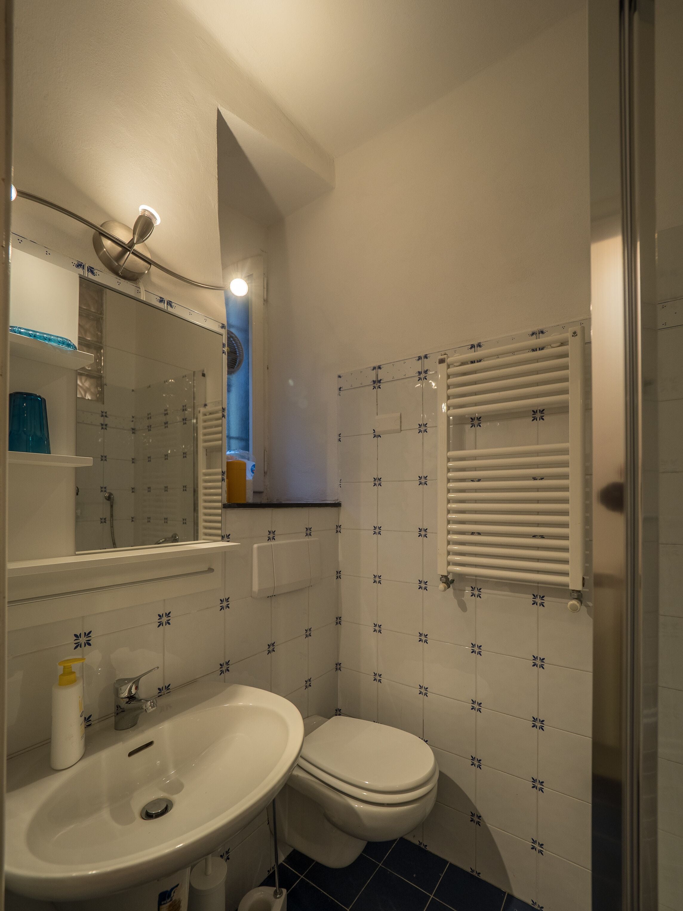 Un baño con lavamanos, inodoro y espejo. Hay una ventana con persianas y una zona de ducha.
