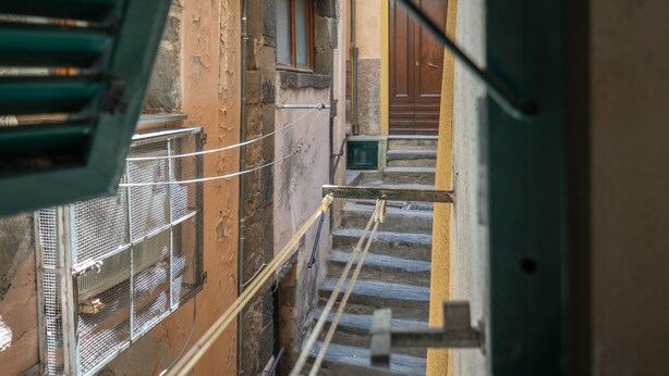 Una escalera estrecha que lleva a una puerta de madera, con barandilla metálica a un lado y fachada de edificio con pintura descascarada y una ventana.