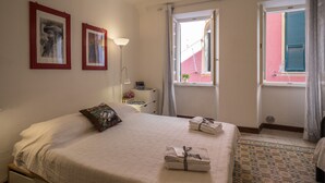 Una cama bien hecha con un cojín, una mesita de noche con una lámpara, y una ventana con vistas al exterior.