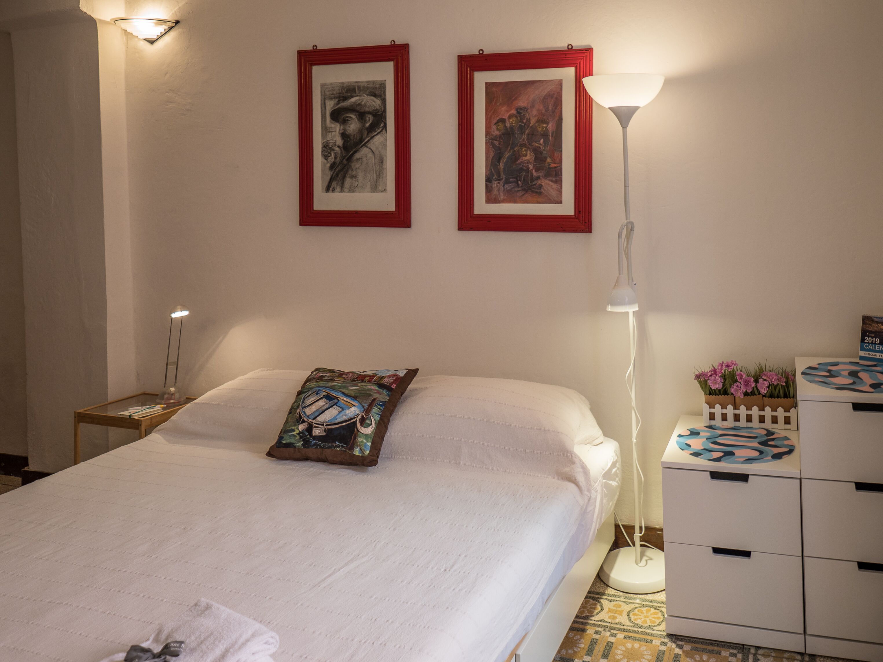 A neatly arranged bedroom with a bed, two framed artworks, a bedside lamp, a nightstand, and a dresser with decorative items.