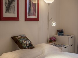 A bedroom with a bed, a nightstand with a calendar, a lamp, and two framed artworks on the wall.
