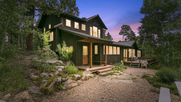 Une maison en bois à deux étages, dotée d’un porche et d’un espace extérieur aménagé pour s’asseoir, le tout entouré d’un paysage naturel.