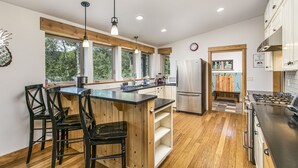 Une cuisine moderne agrémentée d’éléments en bois, avec un îlot central doté de tabourets, des appareils en acier inoxydable et de grandes fenêtres offrant une vue sur la verdure.