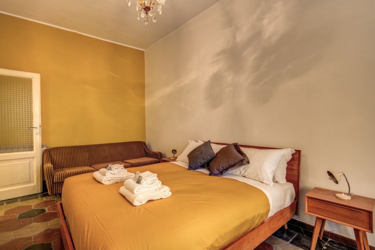 A hotel room with a bed, a bench, a bedside table, and a chandelier.
