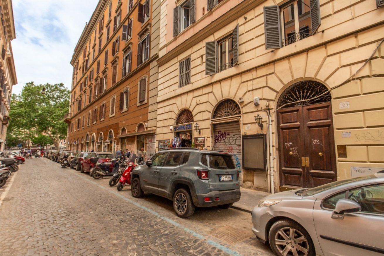 A cobblestone street lined with parked cars and motorcycles, buildings with arched windows and shutters, and a sign for a store.