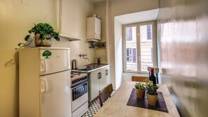 A compact kitchen with a refrigerator, stove, and sink. There is a dining table with chairs and a potted plant. A window provides natural light and a view of buildings outside. - Trastevere Vintage (Rome)