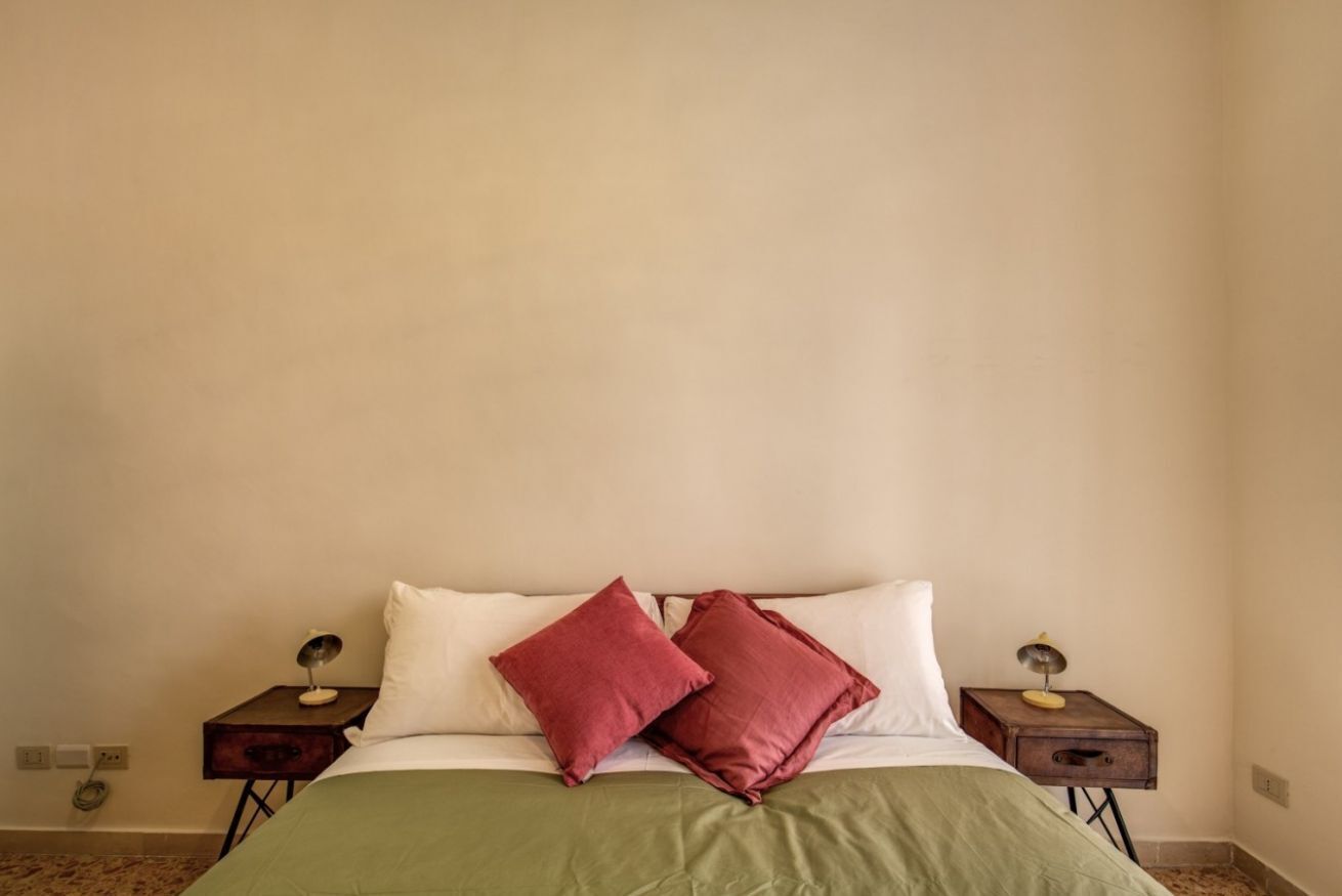Una cama con una colcha verde, dos almohadas blancas y dos almohadas rojas. Hay dos mesitas de noche de madera, cada una con una lámpara a los lados de la cama.