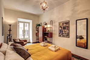A hotel room with a bed, a chair, a bookshelf, and a chandelier.