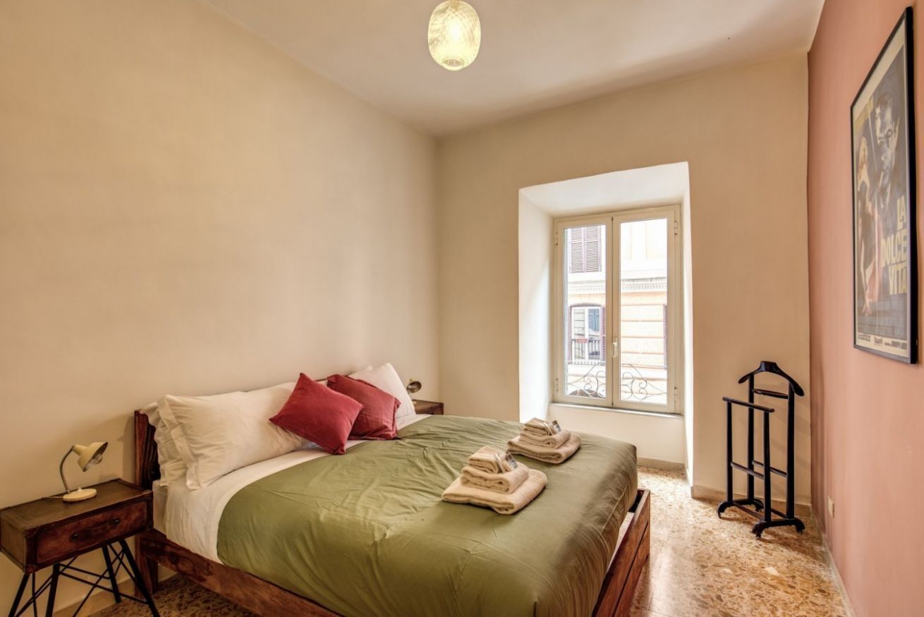 A bedroom with a bed, a nightstand, a window, and a wall-mounted rack.