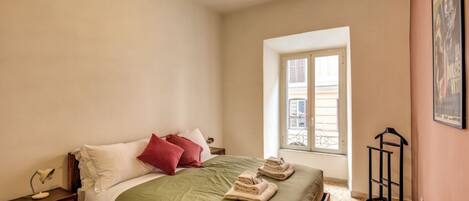 A bedroom with a bed, a nightstand, a window, and a wall-mounted rack.
