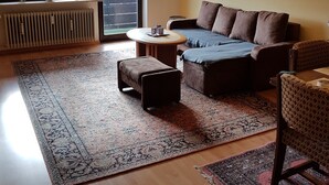 Una sala de estar con un sofá, un reposapiés y una mesa redonda de centro. Hay un radiador, una ventana con persianas y un cuadro en la pared.