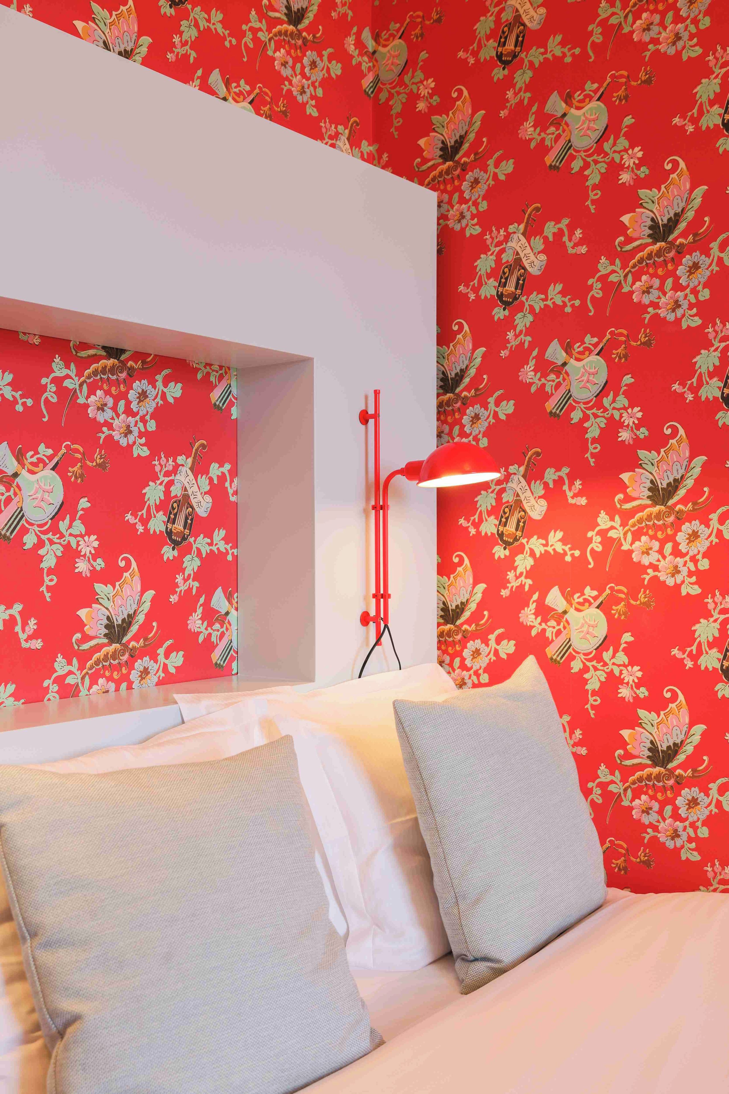 Una habitación con cama, almohadas, una lámpara roja y un papel tapiz floral en la pared.