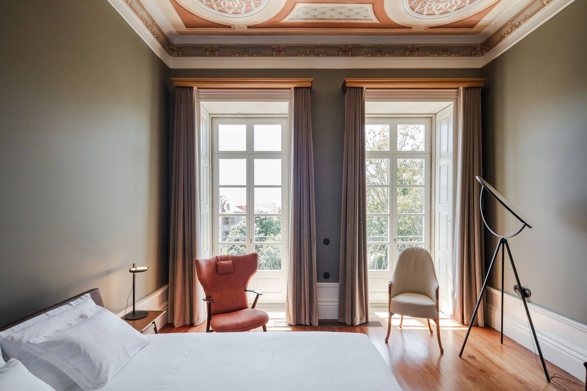 A hotel room with a large bed, two chairs, a tripod, and a window with curtains.
