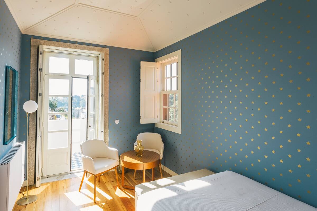 A room with a bed, two chairs, a small table, and a window with star-patterned wallpaper.