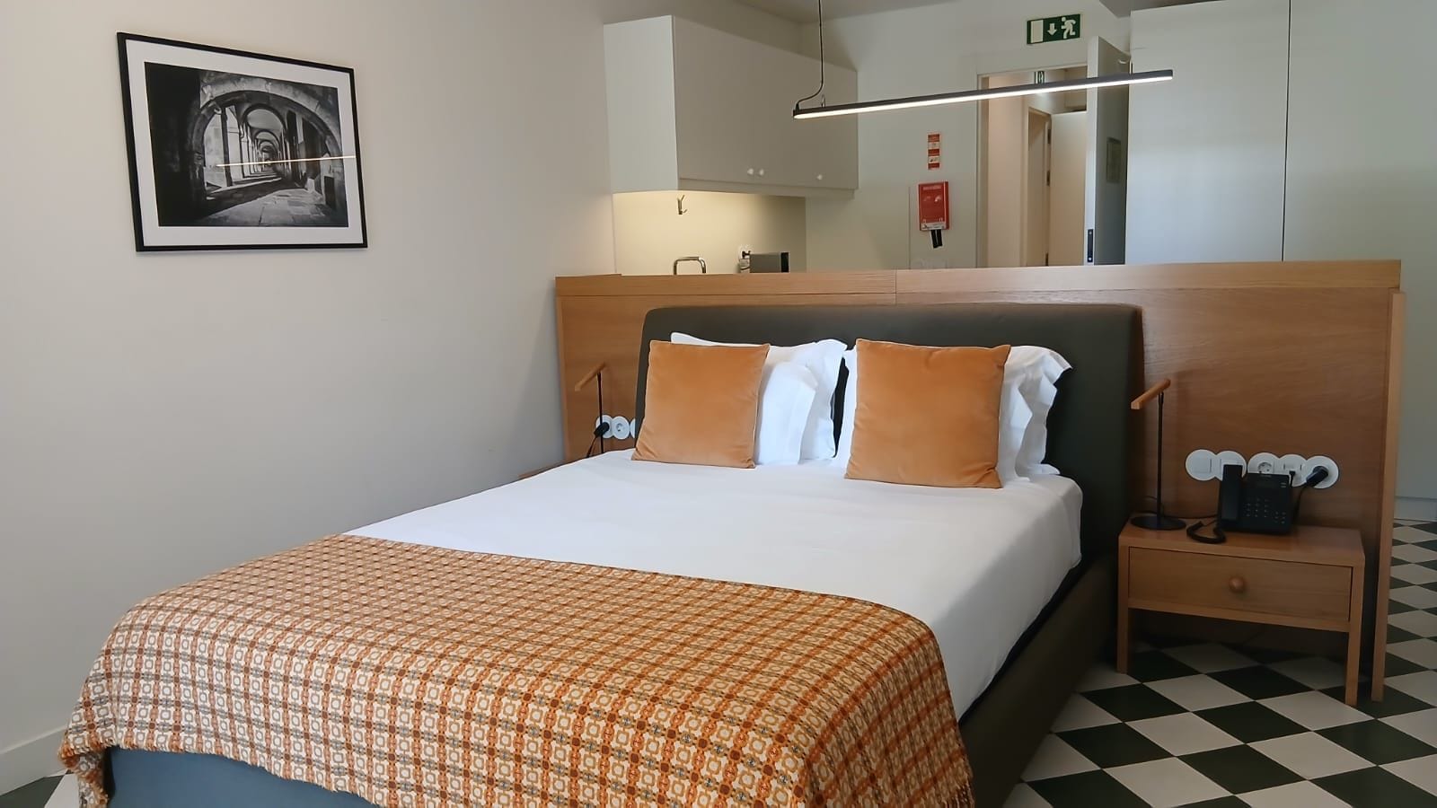 A hotel room with a bed, a nightstand, a lamp, a framed picture, and a checkered floor.