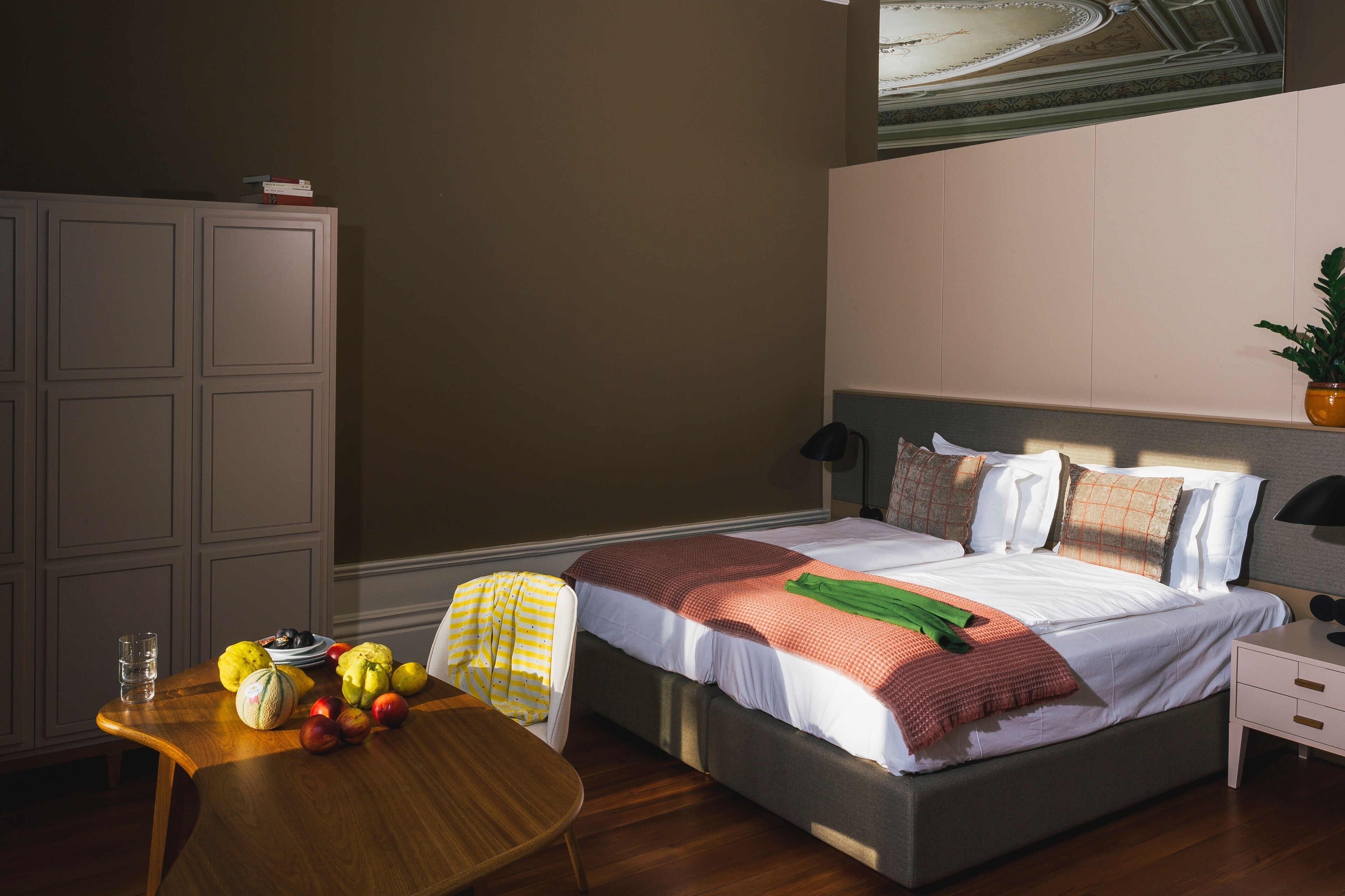 Quarto com cama, mesa de cabeceira e uma cadeira de madeira. Há um guarda-roupa e uma planta na parede.