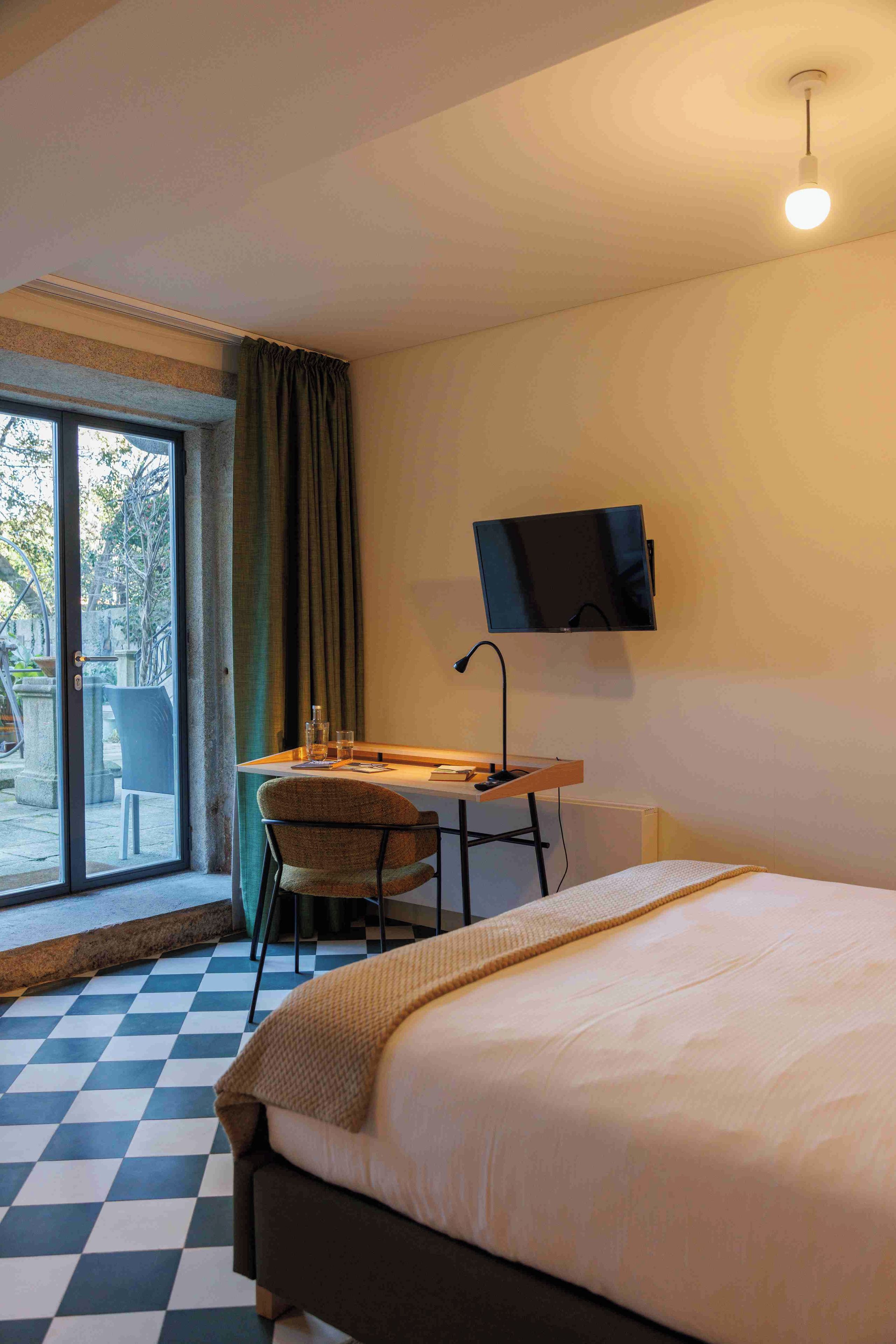 Habitación de hotel con cama, escritorio con silla, televisor montado en la pared y una ventana grande con cortinas.