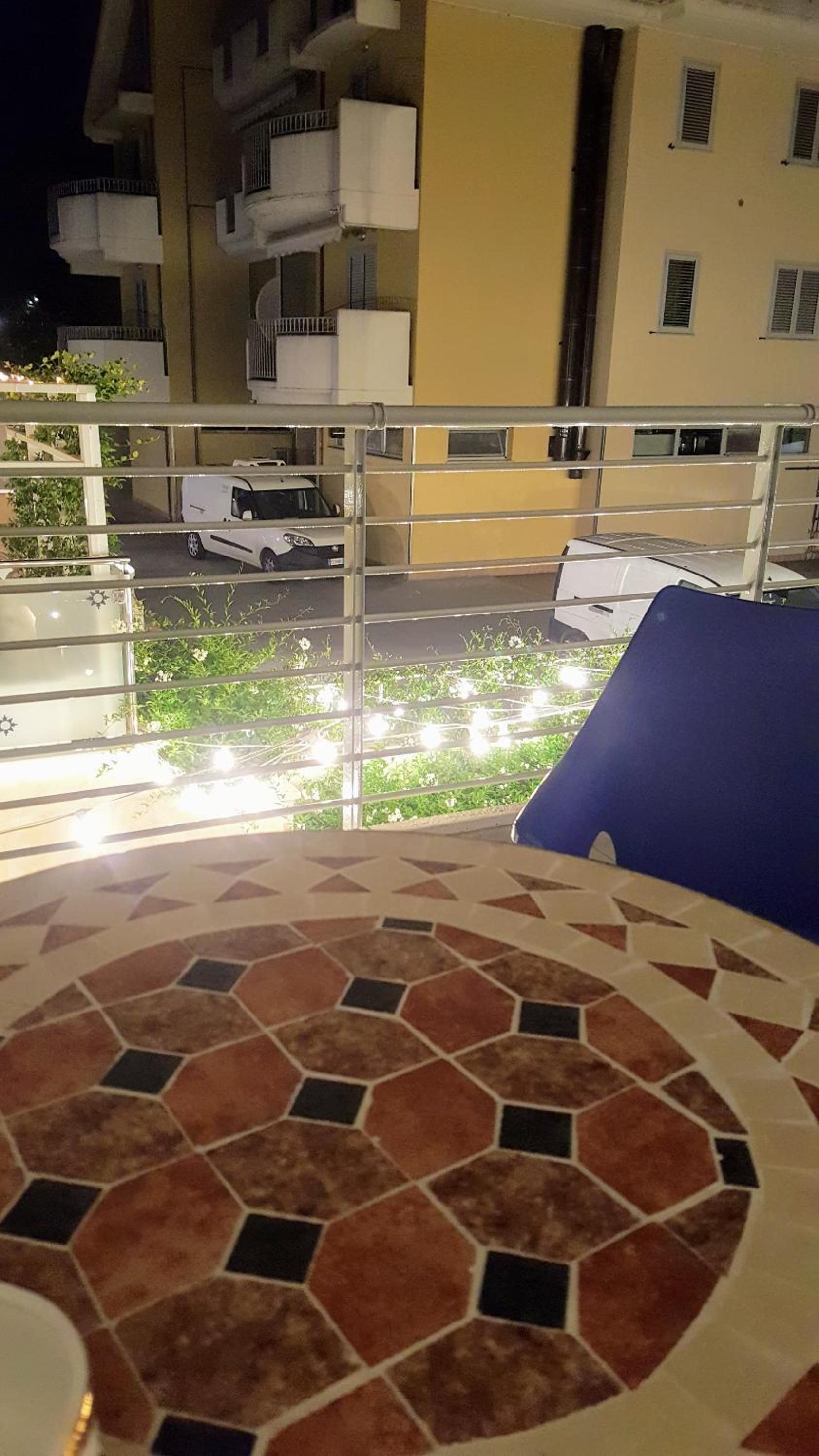 A balcony with a tiled floor, a railing, and a view of a street with parked cars and greenery.