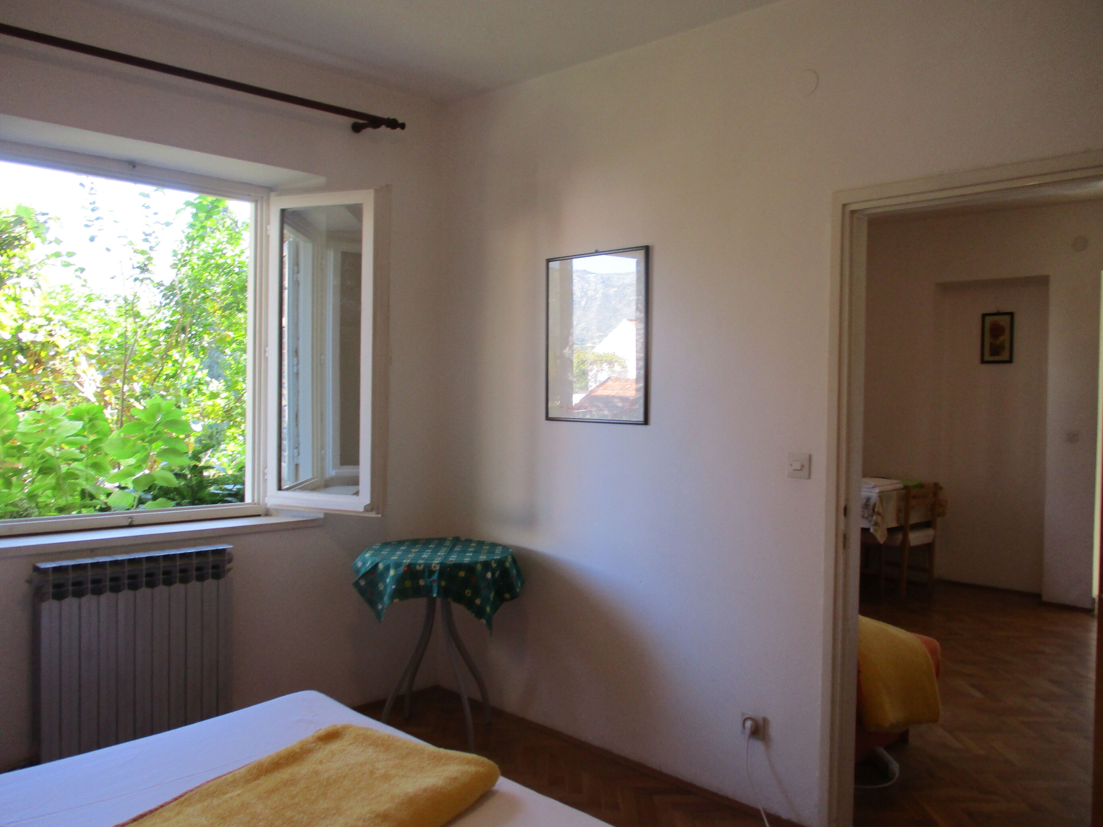 Une chambre avec un lit, un radiateur, une fenêtre donnant sur un espace verdoyant, une petite table recouverte d’une nappe et un tableau encadré accroché au mur.