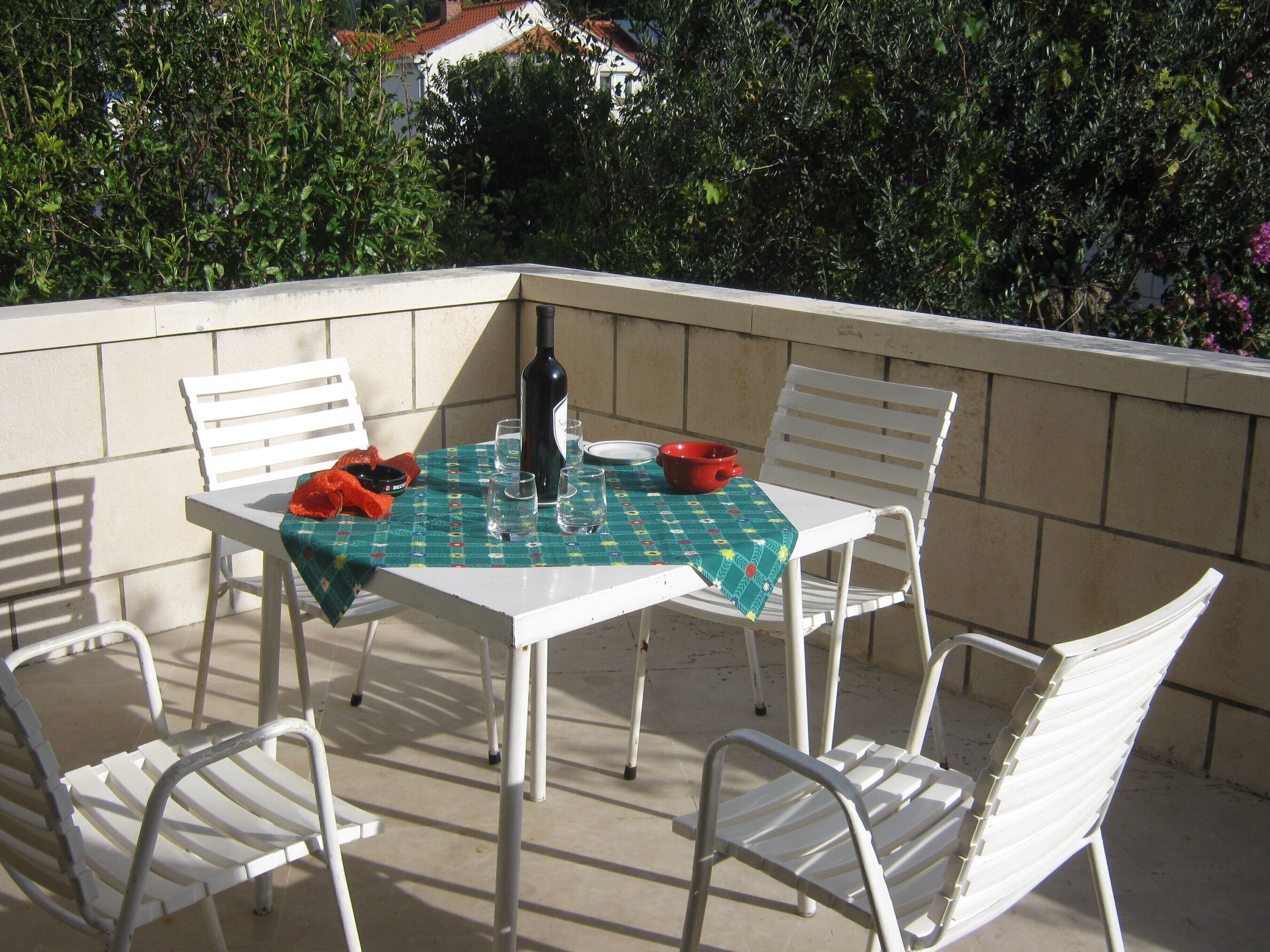 Patio esterno con tavolo apparecchiato per quattro, circondato da sedie bianche, una bottiglia di vino e una ciotola rossa.