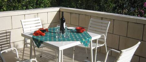 Patio esterno con tavolo apparecchiato per quattro, circondato da sedie bianche, una bottiglia di vino e una ciotola rossa.