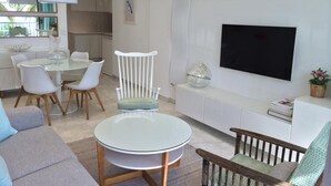 A modern living room with a round glass table, white chairs, a grey sofa, a wooden chair, a TV, and a kitchen in the background. -  CHARMING APARTMENT ON THE BEAUTIFUL PROMENADE OF ENGLISH FACING THE SEA AND BEACHES (Nice)
