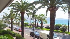 A street lined with palm trees and parked cars, leading to a beach with the ocean in the background. -  CHARMING APARTMENT ON THE BEAUTIFUL PROMENADE OF ENGLISH FACING THE SEA AND BEACHES (Nice)