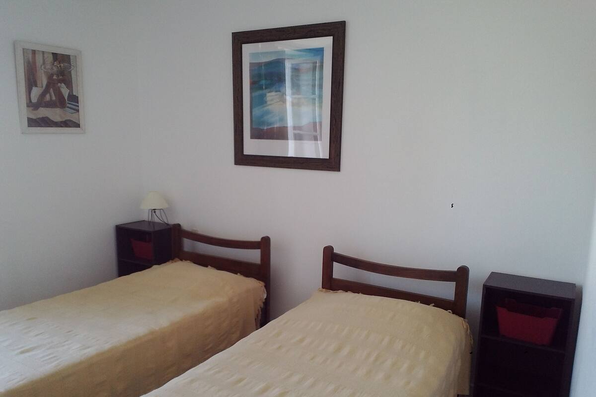 A room with two single beds, a nightstand with a lamp, a framed picture on the wall, and a small cabinet.