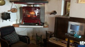 A cozy living room with a wood-burning stove, a comfortable armchair, a small table, and a television on a stand. - La Coquille Napoléon (Uhart-Cize)