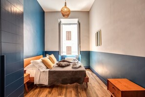 Un dormitorio con cama, mesitas de noche de madera, lámpara colgante y vista a un edificio a través de una ventana.