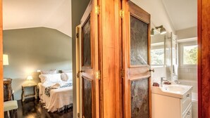 Un dormitorio con un armario de madera, una cama con ropa de cama blanca, una mesita de noche con una lámpara y vistas al baño con lavabo y espejo.