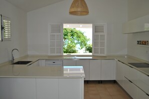 Una cocina moderna con armarios blancos, lavavajillas integrado, un amplio ventanal con contraventanas y una lámpara colgante.