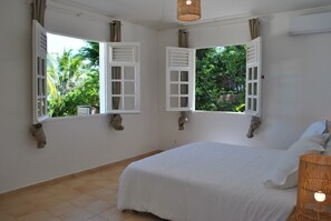 Un dormitorio con cama, dos ventanas con contraventanas, una lámpara colgante y vistas a la vegetación exterior.