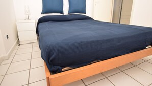 A bed with a wooden frame and a dark blue mattress in a room with white walls and a tiled floor. - Promenade, overlooking the Marciana Marina promenade (Marciana Marina)