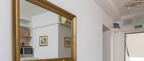 A room with a dining table, a vase with yellow flowers, a bottle of wine, and a glass. There is a large mirror on the wall and a white door with an exit sign.