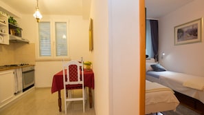 Eine kleine Küche mit Esstisch und Stuhl, ein Bett mit einzelner Matratze und ein kleines Badezimmer, das durch eine geöffnete Tür sichtbar ist.