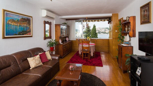 A living room with a brown leather sofa, a wooden coffee table, a red rug, a dining table with chairs, and a TV stand. - Apartments Cime (Dubrovnik)