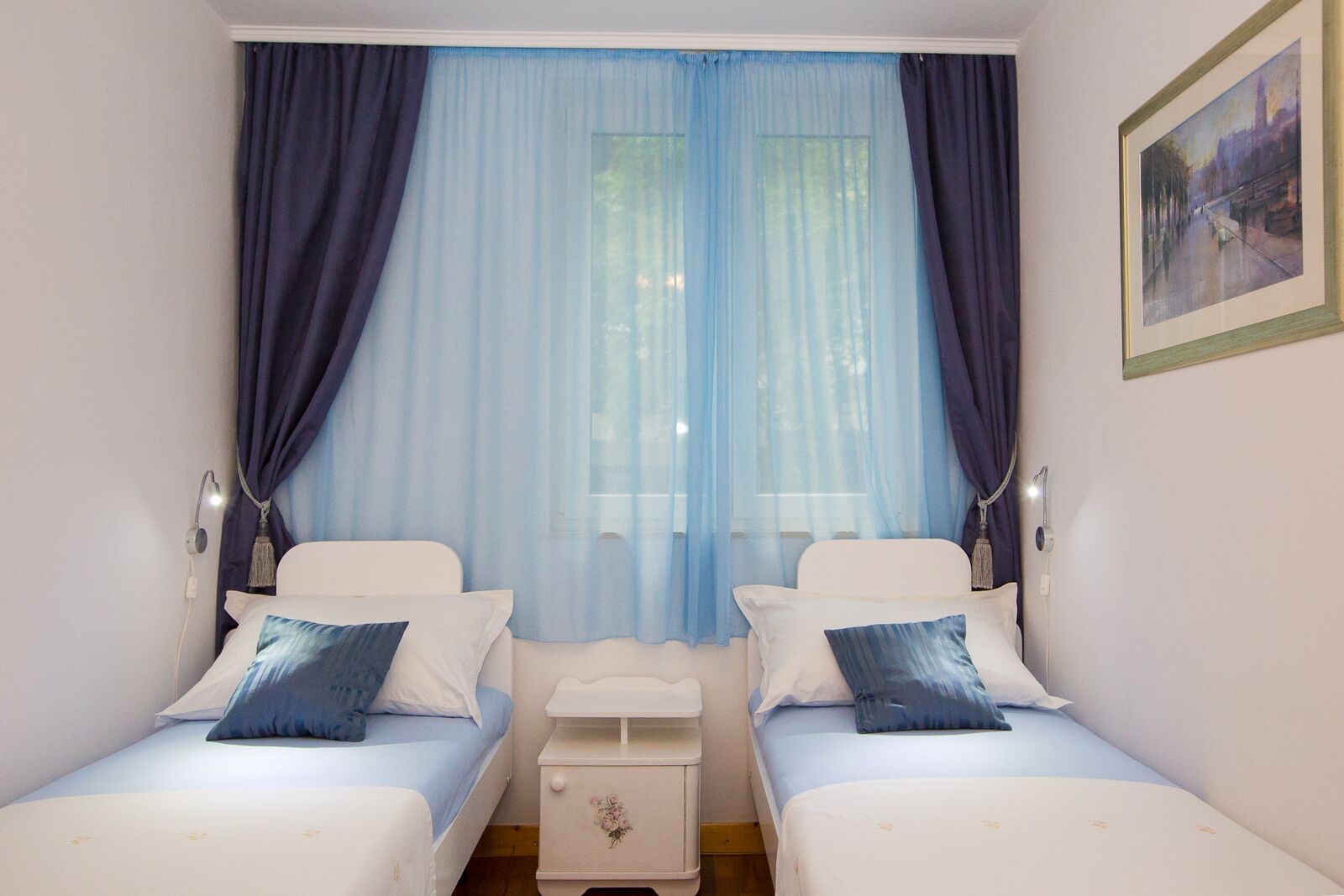 Un dormitorio con dos camas individuales, una mesita de noche, una lámpara y una ventana con cortinas translúcidas.