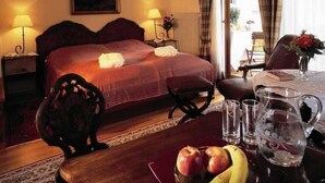 Une chambre d’hôtel avec un grand lit, deux lampes de chevet, un lustre, une table à manger avec une carafe et des verres, et une vue sur l’extérieur par la fenêtre.