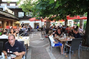 An outdoor dining area with tables and chairs under red umbrellas, surrounded by trees.