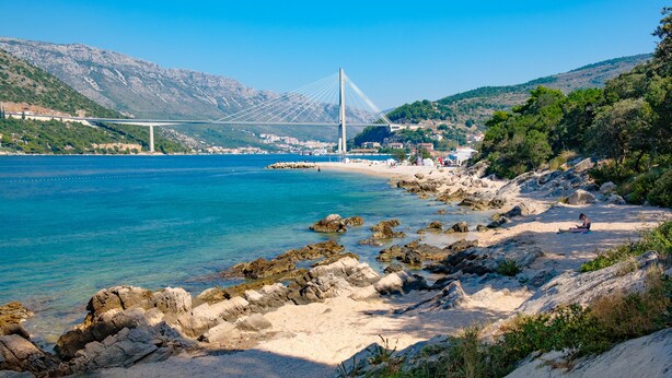 A coastal area with a bridge, rocky beach, and greenery. - Apartment Filip (Dubrovnik)