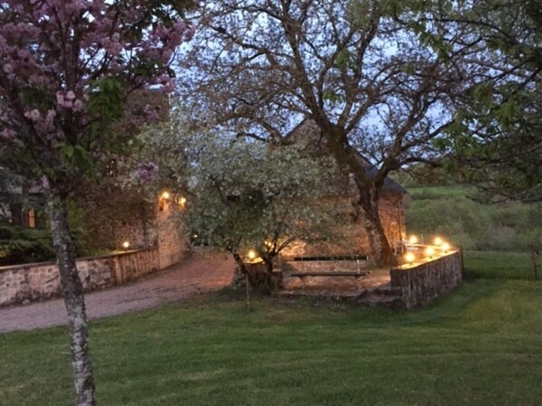 A rustic stone building with a wooden staircase leading up to a door, surrounded by a well-maintained lawn and flowering trees.