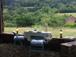 An outdoor dining area with stone walls, two chairs, a table with a tablecloth, and two lit lanterns.