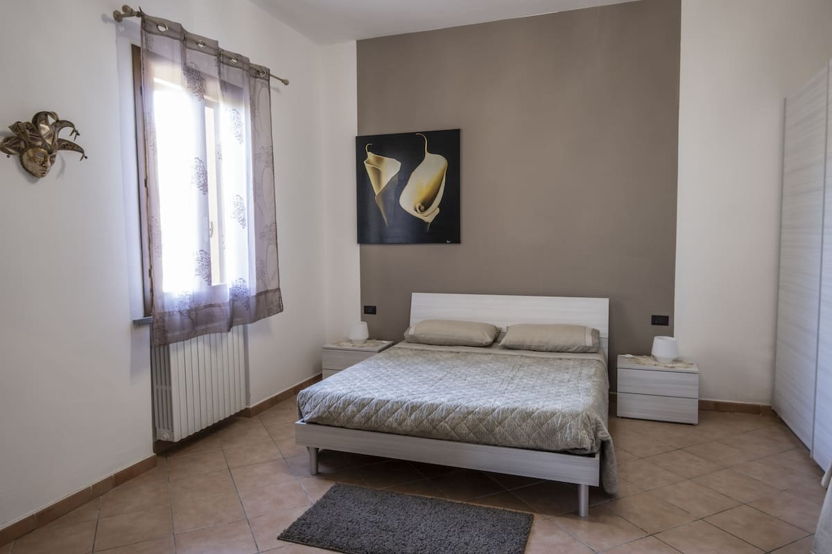Un dormitorio con cama, mesitas de noche, un radiador, una ventana con cortinas y un cuadro colgado en la pared.