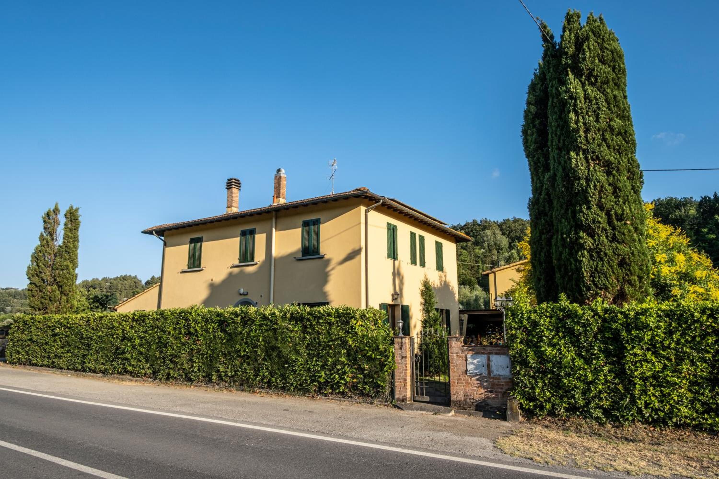 A two-story building with a beige facade, green shutters, and a chimney. It is surrounded by a well-maintained hedge and a tall evergreen tree.