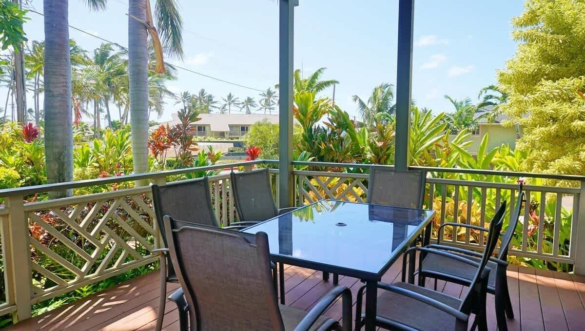 Une terrasse extérieure avec une table et des chaises en verre, entourée d’une clôture en treillis blanc, de palmiers et de plantes tropicales.