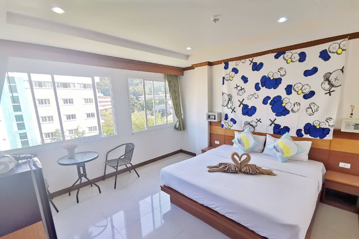 A hotel room with a bed, a small table, a chair, and a view of buildings through large windows.